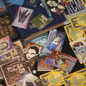 May include: A collection of playing cards and stationery items featuring pixelated graphic illustrations, including portraits, a bottle, and abstract geometric patterns. The items are arranged on a dark surface, with a blue binder and a pen visible.