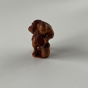 May include: A small, intricately carved wooden figurine depicting a person bent over, carrying a basket and a bundle on their back. The figure is a warm brown color, with detailed carving of clothing and facial features. The figurine is set against a plain white background.