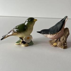 May include: Two ceramic bird figurines. One is green and tan, perched on a white flower. The other is gray, black, and pink, perched on a brown tree branch. Both have detailed features and are set against a white background.