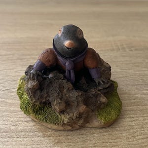 May include: A figurine of a mole wearing a purple scarf and a brown jacket. The mole is sitting on a small, textured base with green and brown details. The mole has a black head and a pink nose.