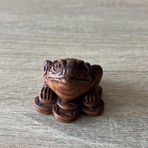 Op de afbeelding: Een gedetailleerd houten kikkerbeeldje, gesneden met ingewikkelde details. De kikker is bruin en lijkt op een stapel munten te zitten. Het beeldje is een decoratief item, mogelijk voor een bureau of plank.