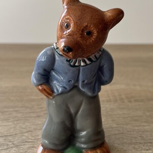 May include: A ceramic figurine of a bear wearing a blue jacket, gray pants, and brown shoes. The bear has a brown head with black eyes and a white collar. The figurine is standing on a small green base.