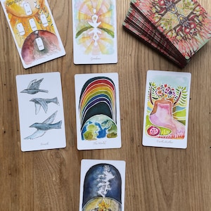May include: A spread of tarot cards on a wooden table. The cards are from the Garden Spread deck and include cards like "Goodness", "The World", "Earth Mother", "Breath", and "Fire".