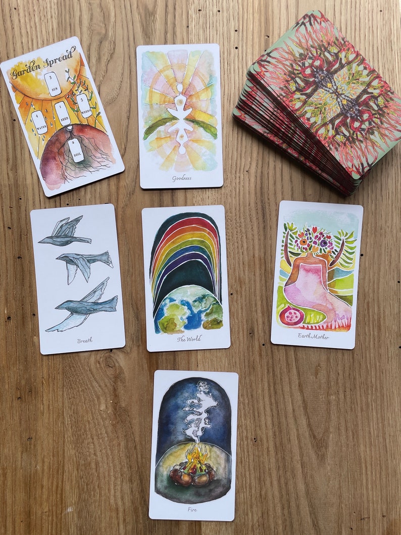 May include: A spread of tarot cards on a wooden table. The cards are from the Garden Spread deck and include cards like "Goodness", "The World", "Earth Mother", "Breath", and "Fire".