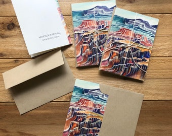 May include: A set of five watercolour note cards featuring a landscape scene of a canyon with red, orange, and purple hues. The cards are tied together with twine and are packaged with brown envelopes.
