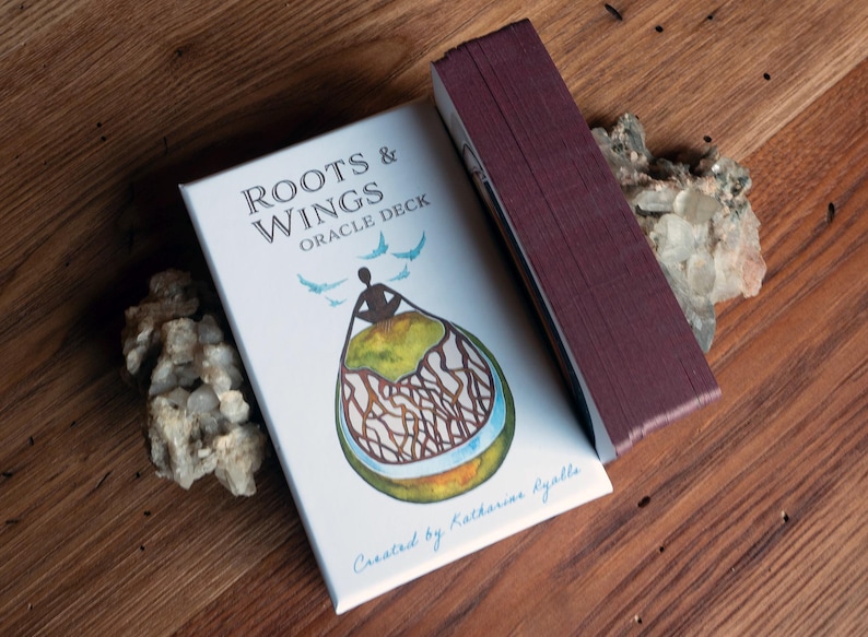 May include: A white box with the text "Roots & Wings Oracle Deck" and an illustration of a person sitting on a green island with roots and wings. The box is on a wooden surface with a pile of white crystals.