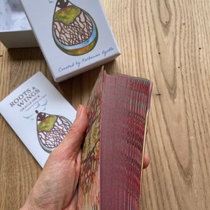 May include: A deck of Roots & Wings Oracle Cards, created by Katherine Gallagher. The cards are stacked and have a burgundy back with a gold and green design. The box is white with a blue and green illustration of a person with roots and wings.