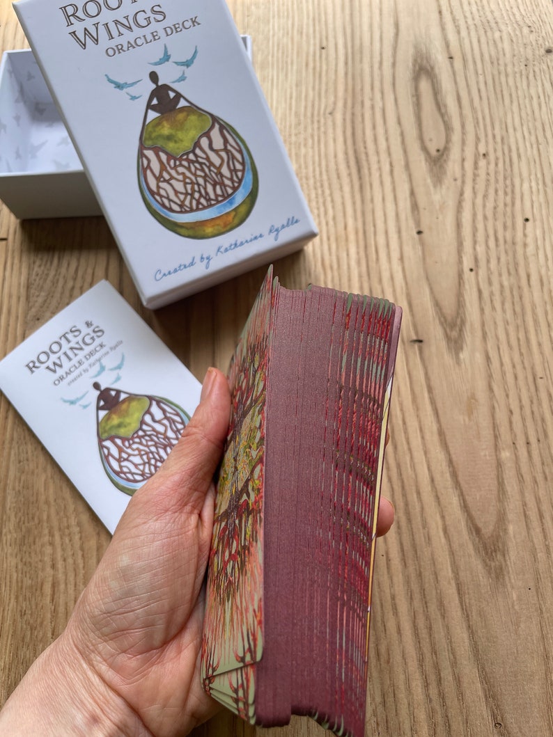 May include: A deck of Roots & Wings Oracle Cards, created by Katherine Gallagher. The cards are stacked and have a burgundy back with a gold and green design. The box is white with a blue and green illustration of a person with roots and wings.