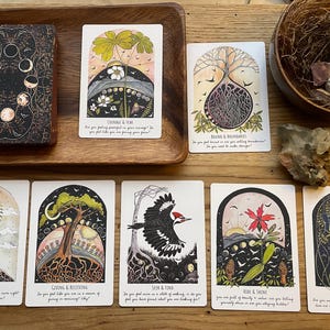 May include: A deck of tarot cards with black and gold moon phase illustrations, alongside seven individual cards featuring nature-themed artwork, including trees, birds, and flowers, with printed text captions below each illustration on a wooden surface.