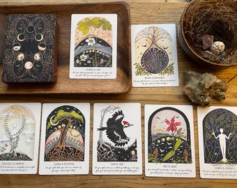 May include: A deck of tarot cards with black and gold moon phase illustrations, alongside seven individual cards featuring nature-themed artwork, including trees, birds, and flowers, with printed text captions below each illustration on a wooden surface.