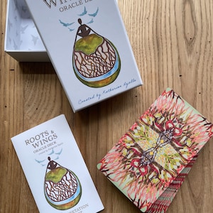 May include: A white box with the text "Roots & Wings Oracle Deck" and a colorful illustration of a person sitting on a tree root with birds flying above. The box is open and the deck of cards is visible. The cards have a colorful, abstract design.