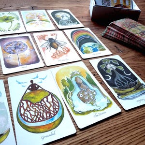 May include: A set of tarot cards with colorful illustrations and text. The cards feature images of nature, animals, and people. The cards are laid out on a wooden table.