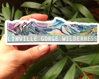 May include: A sticker with a watercolor painting of a mountain range in shades of blue, green, and purple. The text "Linville Gorge Wilderness" is printed in white on a teal background at the bottom of the sticker.