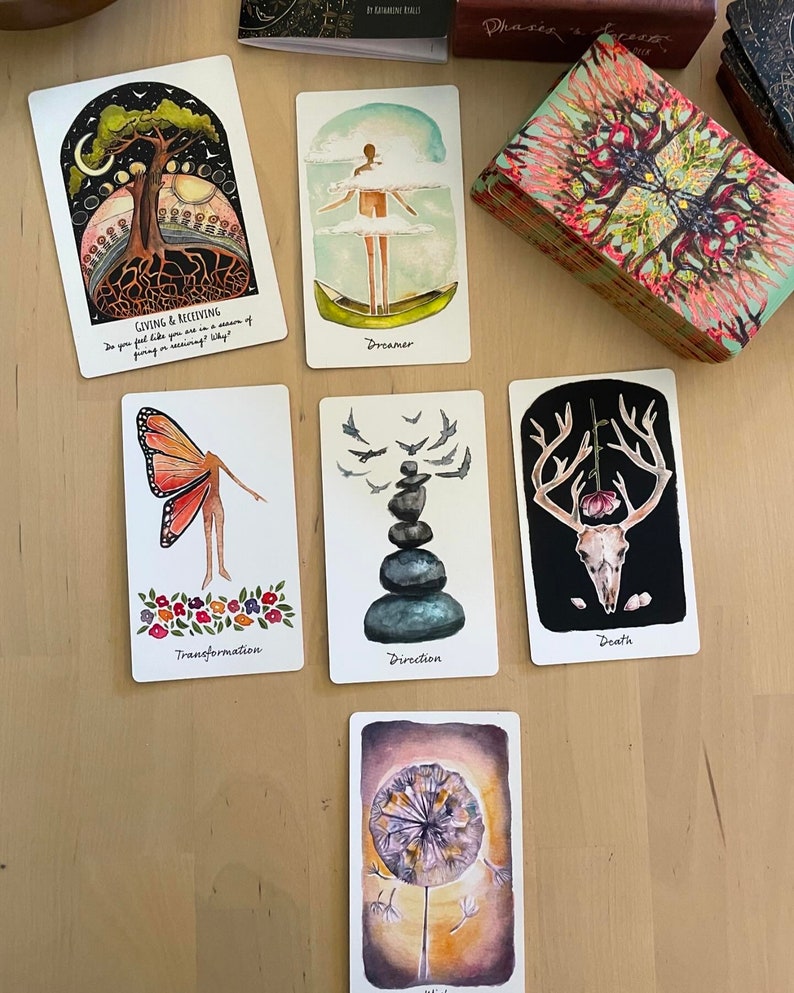 May include: A set of tarot cards with colorful illustrations. The cards feature images of a tree with roots, a person in a boat, a butterfly, a stack of stones, a deer skull, and a dandelion.