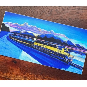 May include: A yellow and black train engine with the word "ALASKA" and the number "4319" on the side, traveling through a snowy landscape with mountains in the background.