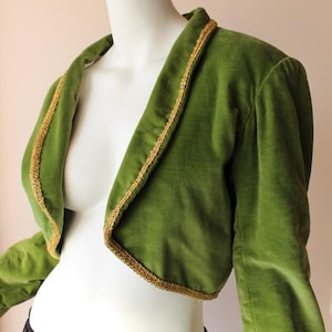 Puede incluir: Chaqueta bolero de terciopelo verde con ribete dorado. La chaqueta de manga corta tiene cuello en V y se muestra en un maniquí. El ribete dorado acentúa los bordes de la chaqueta, añadiendo un toque de elegancia. La chaqueta es de un tono verde vibrante.