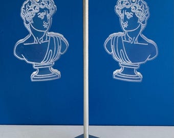 May include: A pair of clear acrylic earrings featuring a white outline of a bust sculpture. The earrings are hanging from silver hooks on a silver stand.