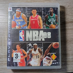 Pode incluir: Capa de jogo de vídeo PlayStation 3 para NBA 08. A capa apresenta imagens de jogadores de basquete em ação, com o logotipo da NBA em destaque. A capa do jogo está em uma caixa de plástico transparente.