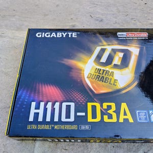 Könnte beinhalten: Eine schwarze und gelbe Schachtel für ein GIGABYTE H110-D3A Ultra Durable Motherboard. Die Schachtel zeigt das GIGABYTE-Logo, die Worte „ULTRA DURABLE“ und die Intel Core- und Chipsatz-Logos. Die Schachtel liegt auf einer grauen Oberfläche.
