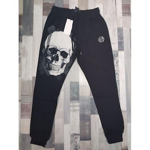 May include: Black sweatpants with a large skull graphic on the left leg. The skull is white with black shading. The trousers have a drawstring waist and a small logo on the right leg.
