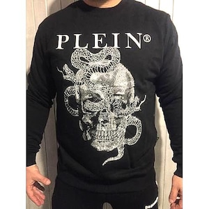Op de afbeelding: Zwart sweatshirt met lange mouwen en een witte afbeelding van een schedel met slangen. Het woord "PLEIN" staat in het wit bovenaan. De sweatshirt is gemaakt van een zacht, comfortabel materiaal.
