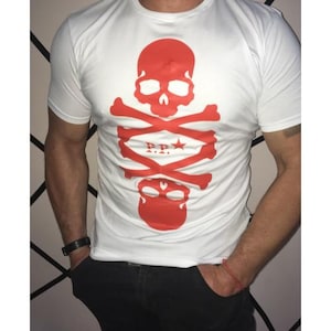 Könnte beinhalten: Weißes T-Shirt mit einem auffälligen roten Totenkopf- und gekreuzten Knochen-Design. Das Design enthält einen Stern und die Buchstaben "P.P.". Das kurzärmelige Shirt ist aus weichem Material. Das Design ist mittig auf der Vorderseite.