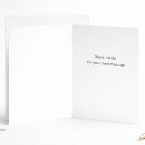 May include: A white folded greeting card standing upright, featuring the black printed text "Blank inside for your own message" on the right interior panel. The card is displayed with a gold pen and small metal paper clips nearby.