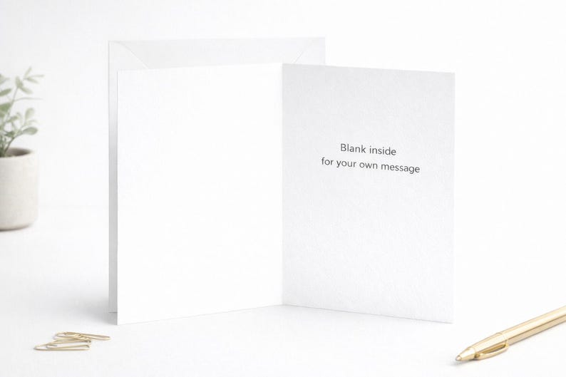 May include: A white folded greeting card standing upright, featuring the black printed text "Blank inside for your own message" on the right interior panel. The card is displayed with a gold pen and small metal paper clips nearby.