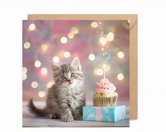 May include: A square greeting card featuring a fluffy gray tabby kitten sitting next to a cupcake with pink frosting and a single lit candle, placed on a light blue gift box, set against a background of soft, out-of-focus bokeh lights.