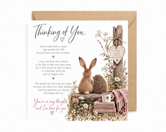 May include: A square sympathy greeting card with a brown envelope. The card features a rabbit and hedgehog sitting on a wooden block, a mug, stacked books, and wildflowers. Text reads: Thinking of You, Always Loved Never Forgotten, and You're in my thoughts and I'm here for you.