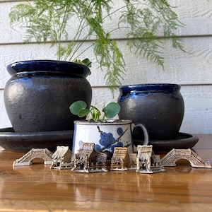 May include: Two dark charcoal ceramic planters with deep blue rims, a white ceramic mug featuring a blue floral pattern, and a collection of small, detailed miniature clay structures including houses and arched bridges arranged in a row.