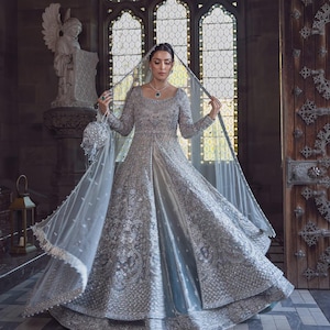 May include: A floor-length silver gown with long sleeves, a scoop neckline, and intricate embroidery throughout. The dress features a front slit, a full skirt, and a matching sheer veil with beaded edges. A small, round, silver beaded handbag is held.