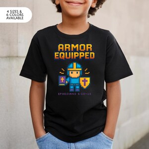 Op de afbeelding: Zwart t-shirt met de tekst "ARMOR EQUIPPED" in gele en blauwe pixellettertype. Een gepixeleerde cartoonfiguur in blauw harnas houdt een boek en schild vast. De tekst "Efeziërs 6:10-18" staat eronder. Verkrijgbaar in 4 maten en 6 kleuren.