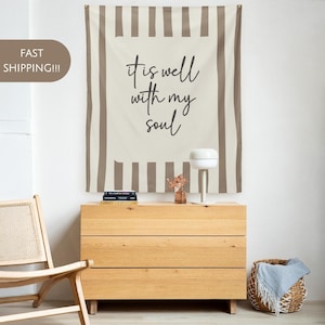 Puede incluir: Un tapiz de pared a rayas beige y marrón con la frase "it is well with my soul" en escritura negra. El tapiz cuelga sobre una cómoda de madera con tres cajones. También se ven una lámpara blanca y una cesta de mimbre.