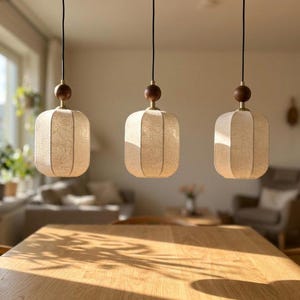 May include: Three pendant lights hanging in a row, each featuring a rectangular, off-white textured shade with rounded corners. Each light is suspended by a black cord and includes a dark brown wooden sphere accent above the shade with brass hardware.
