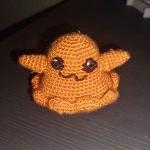 May include: A crocheted orange plush toy with black eyes and a smiling mouth. The toy is sitting on a dark brown surface.