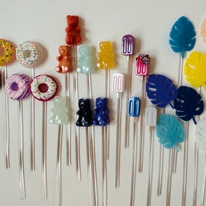 May include: A collection of colorful drink stirrers featuring donuts, gummy bears, and popsicle designs. The stirrers are clear with various colors, including yellow, pink, red, and blue. The overall theme is fun and playful.