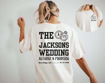 May include: Cream-colored t-shirt with black text and graphics. The back of the shirt features the words "THE JACKSONS WEDDING" with the couple's initials inside of rings. The wedding date is 05.02.2026.