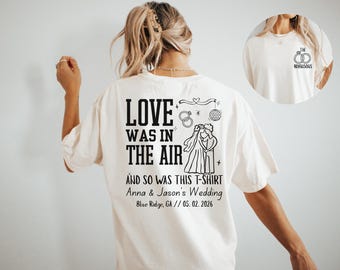 May include: Cream-colored t-shirt with black text and graphics. The back of the shirt reads "LOVE WAS IN THE AIR" with a wedding-themed illustration and the names "Anna & Jason's Wedding" and the date 05.02.2026. A smaller image of the shirt is in the upper right.