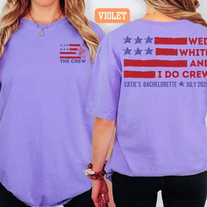 May include: A violet short-sleeved t-shirt with a graphic design on the front and back. The front features a flag design with the text "THE CREW". The back displays a flag design with the text "WED WHITE AND I DO CREW CATIE'S BACHELORETTE JULY 2025".