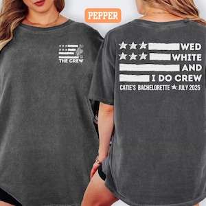 May include: Dark gray t-shirt with a white and gray flag design and the text "THE CREW" on the front. The back of the shirt features the text "WED WHITE AND I DO CREW" and "CATIE'S BACHELORETTE JULY 2025".
