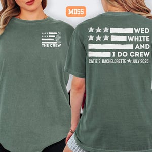 May include: Moss green t-shirts with white text and graphics. The front of the shirt says "THE CREW" with a flag design. The back of the shirt says "WED WHITE AND I DO CREW CATIE'S BACHELORETTE JULY 2025" with a flag design.
