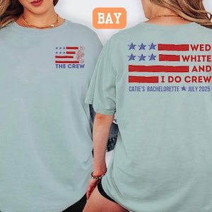 May include: Light blue t-shirt with a red, white, and blue flag design. The front of the shirt says "THE CREW" and the back says "WED WHITE AND I DO CREW CATIE'S BACHELORETTE JULY 2025".