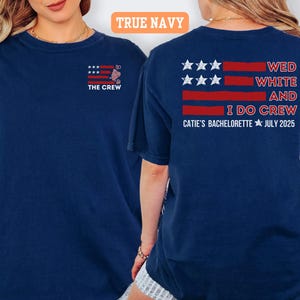 May include: Navy blue t-shirts with a patriotic design. The front of the shirt says "THE CREW" with an American flag graphic. The back of the shirt says "WED WHITE AND I DO CREW CATIE'S BACHELORETTE JULY 2025".