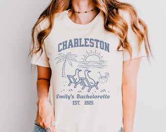 May include: Cream-colored t-shirt with a blue graphic featuring the word "Charleston" above a beach scene with palm trees, sun, and lounge chairs. Below the graphic, it reads "Emily's Bachelorette EST. 2025".