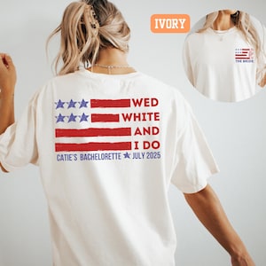 May include: Ivory-colored t-shirt with a patriotic design. The back features a red, white, and blue American flag graphic with the text "WED WHITE AND I DO" and "CATIE'S BACHELORETTE JULY 2025". A smaller flag design is on the front.