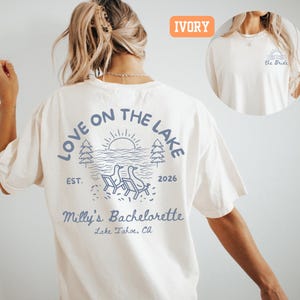 May include: An ivory t-shirt featuring a blue graphic on the back with the text Love on the Lake, Est. 2026, Milly's Bachelorette, and Lake Tahoe, CA. The graphic shows two chairs by a lake with trees and a sun. A small front graphic says the Bride.