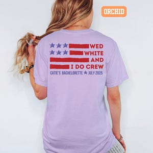 May include: A light purple t-shirt with a red and blue graphic resembling a flag. The text on the shirt reads "WED WHITE AND I DO CREW CATIE'S BACHELORETTE JULY 2025". The shirt is a comfortable, casual style.