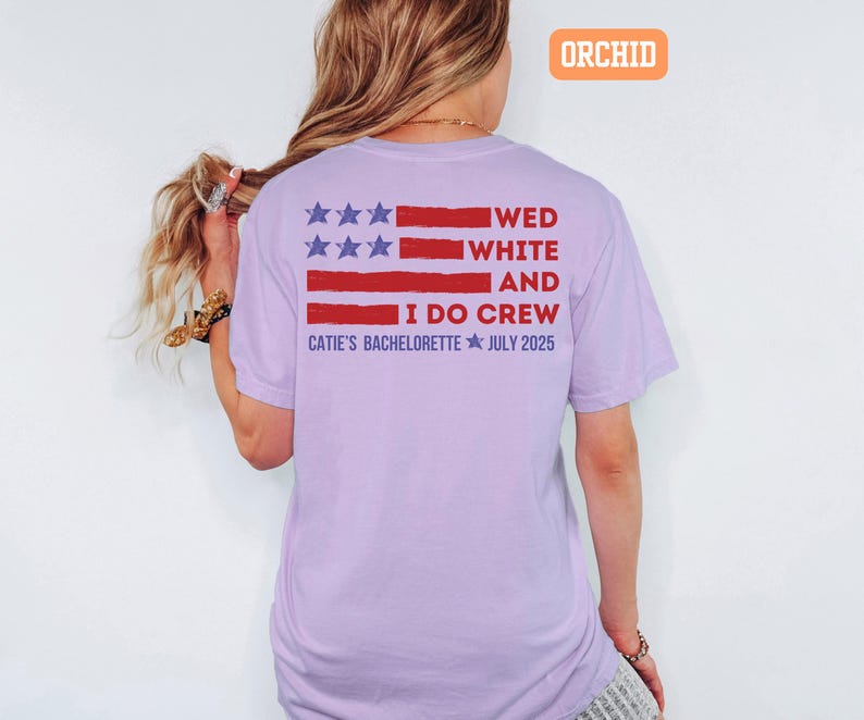 May include: A light purple t-shirt with a red and blue graphic resembling a flag. The text on the shirt reads "WED WHITE AND I DO CREW CATIE'S BACHELORETTE JULY 2025". The shirt is a comfortable, casual style.