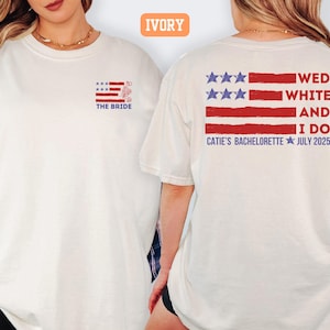 May include: Ivory-colored t-shirt with a patriotic design. The front features a small American flag graphic with the text "THE BRIDE." The back displays a larger flag design with the words "WED WHITE AND I DO" and "CATIE'S BACHELORETTE JULY 2025."